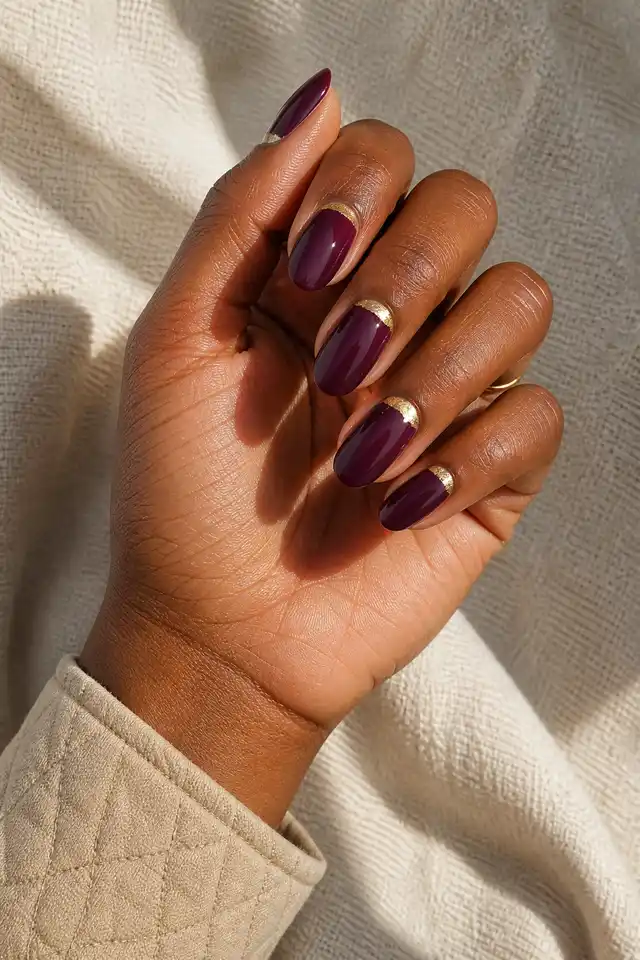 Gold foil crescent accents on deep plum oval New Year's nails with light olive skin and a compact curled pose in natural daylight