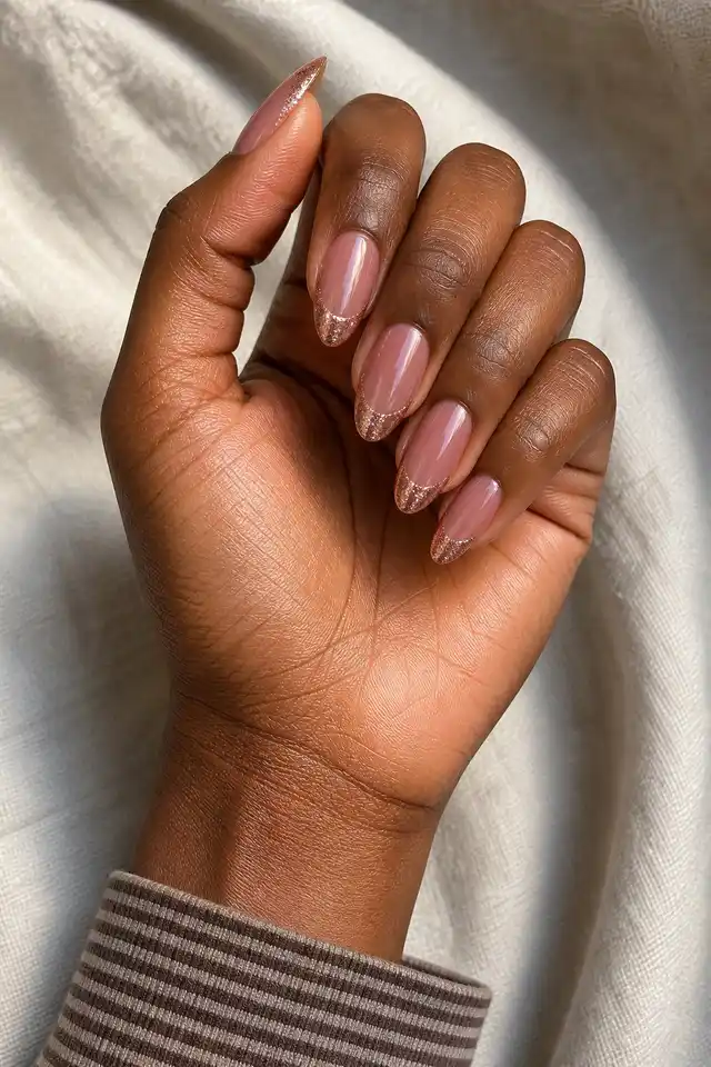 Rose gold sparkle French tips on medium almond nails with rich dark brown skin, a gently curled hand, and a cream sleeve in natural daylight