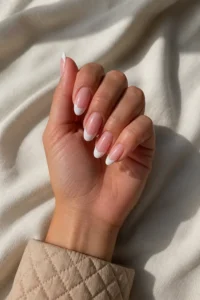 Fair skin hand in a softly curled pose wearing glossy blush-pink almond nails with crisp white French tips and a pale quilted cuff
