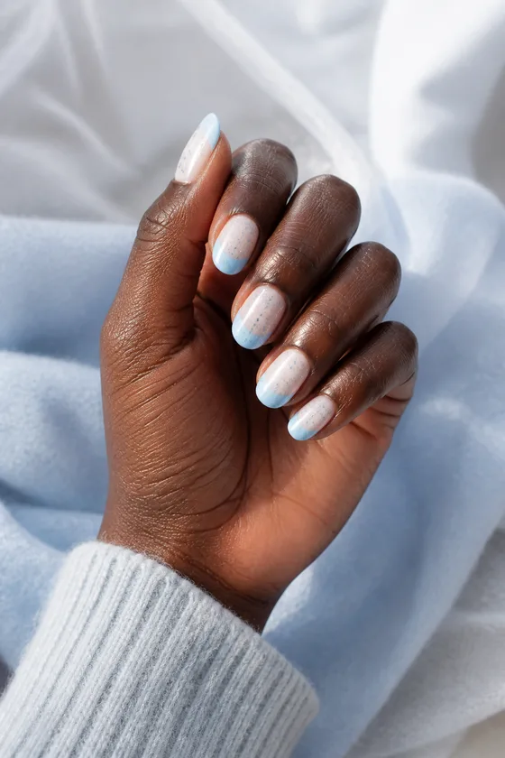 Frosty white short almond nails with pale blue French tips and tiny silver-blue sparkle dots on one deep ebony hand in an upright softly curled pose with a grey-blue cuff, natural daylight