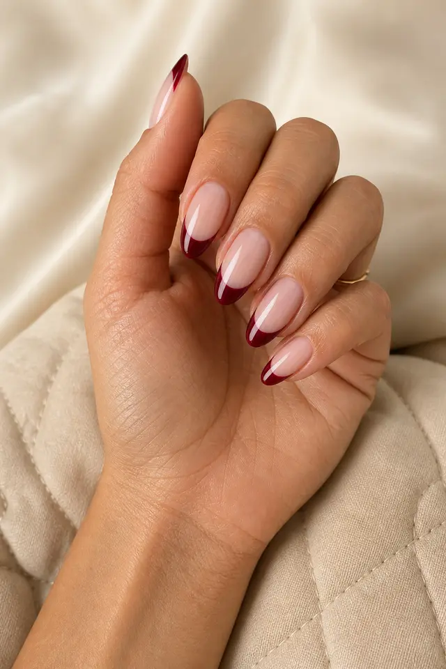 Deep wine French tips on a sheer blush nude base with medium almond nails, a layered fan pose, medium skin, a beige quilted textile, and one tiny gold ring in natural daylight