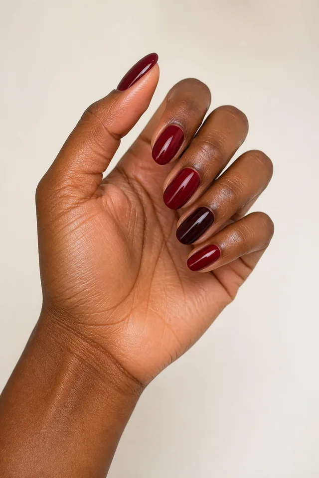 Deep garnet micro French lines on medium oval nails with a curved radial display pose, deep brown skin, and a bare wrist in natural daylight