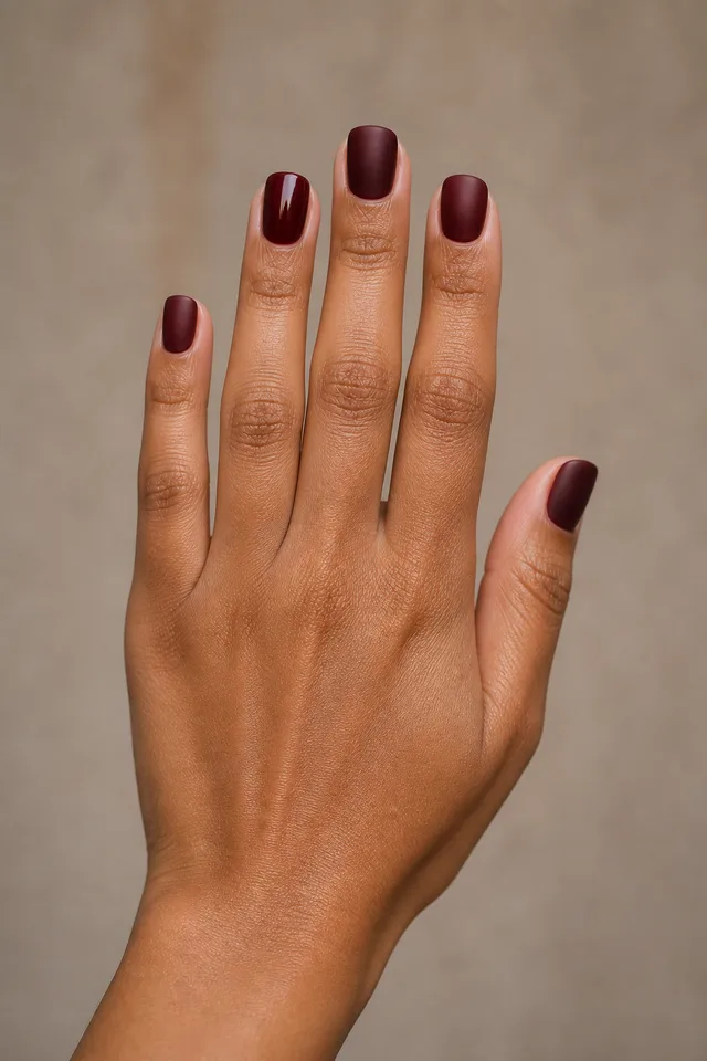 Burgundy satin polish on short squoval nails with an upright narrow fan pose, medium brown skin, and a bare wrist in natural daylight