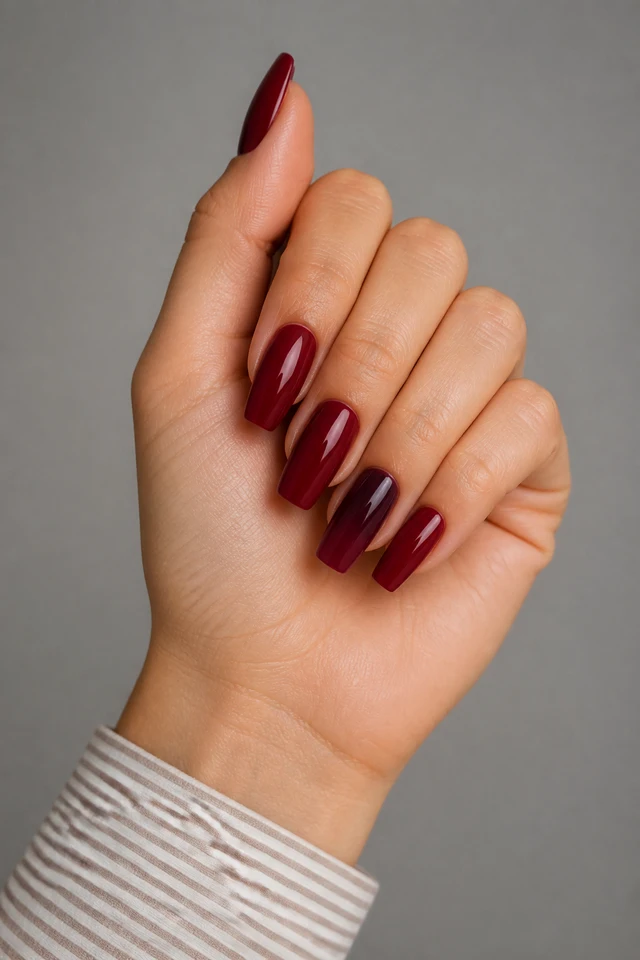 Deep garnet glossy polish on medium ballerina nails with a compact curled fist and raised thumb pose, light olive skin, and a taupe striped sleeve in natural daylight