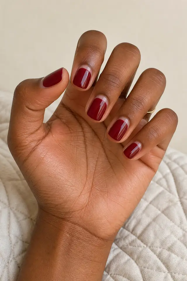 Wine glossy short square nails with sheer cuticle crescents, a layered macro fan pose, deep brown skin, a beige quilted textile, and one tiny gold ring in natural daylight