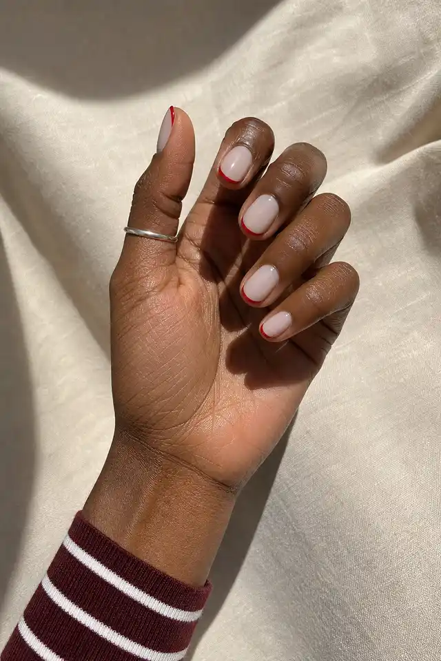 Red micro French tips on short oval nails with a curved radial display pose, rich dark brown skin, and a bare wrist in natural daylight