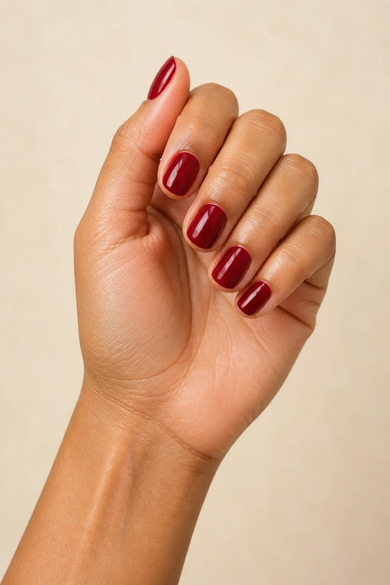 Medium skin hand in a compact softly curled pose wearing short glossy deep cherry red rounded nails against an ivory background
