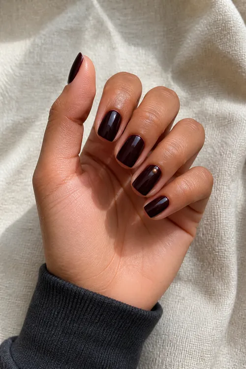 Black cherry short squoval nails with one tiny bronze accent on a tan skin tone hand in a relaxed curled pose with a charcoal sleeve and visible wrist