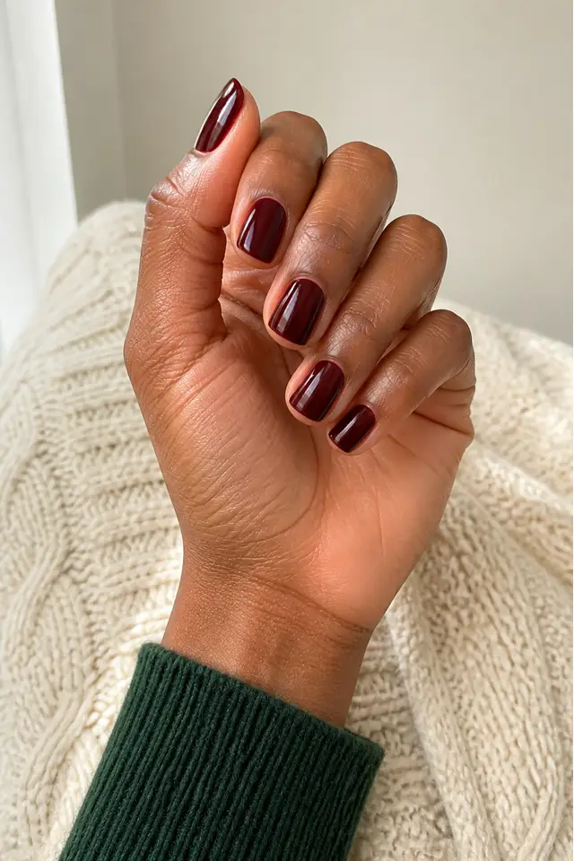 Mulled wine gloss short squoval nails on a softly curled hand with deep golden-brown skin and a forest-green knit cuff in natural daylight